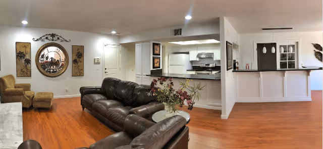 Open-concept common area with leather sofas and armchairs on hardwood floors facing a kitchen with a breakfast bar and recessed lighting.