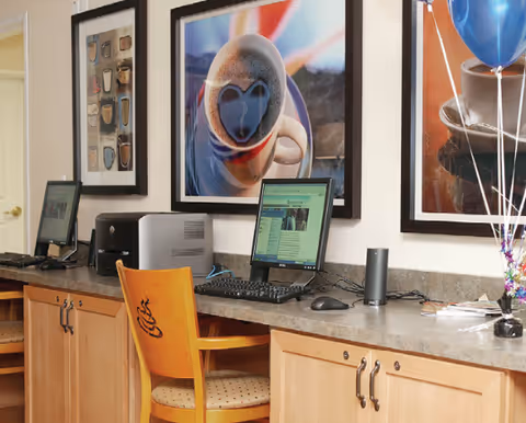 A computer workstation area with two desktop computers on a countertop with wooden cabinets below. A wooden chair with a coffee cup design on the backrest is placed in front of one computer. The wall behind the computers features framed pictures of coffee cups and balloons are visible on the right side.