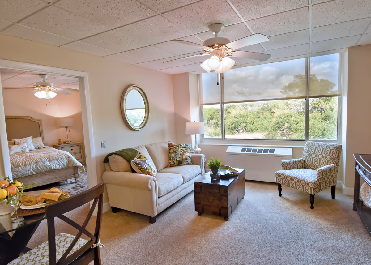 A cozy living room area with a beige sofa adorned with patterned cushions, a wooden chest used as a coffee table, a patterned armchair, and a large window with a view of greenery outside. A round mirror hangs on the wall above the sofa, and a ceiling fan with lights is mounted on the ceiling. To the left, an open doorway reveals a bedroom with a bed, nightstand, lamp, and ceiling fan.
