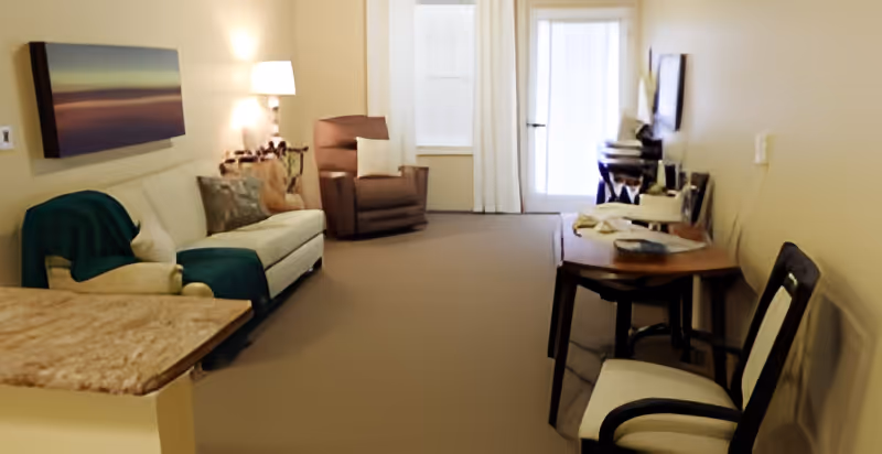 A cozy living room with a white sofa adorned with pillows and a green throw blanket, a brown armchair, a floor lamp, a wall-mounted painting, a wooden table with chairs, and a TV on a stand near a door with white curtains.