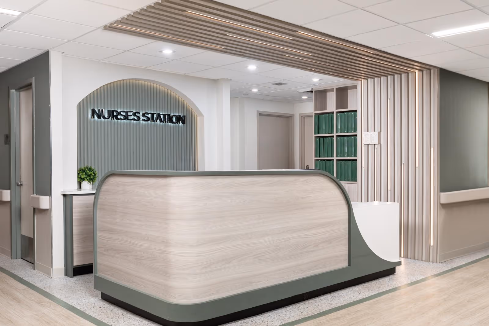A modern nurses station inside a healthcare facility with a curved light wood desk, green accents, a small plant, and a wall sign that reads 'NURSES STATION'. The area is well-lit with ceiling lights and features a clean, organized design with vertical slats on one wall and shelves with green binders.