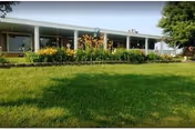 A single-story building with large windows and a covered walkway, surrounded by a well-maintained garden with flowers and green grass under a clear sky.