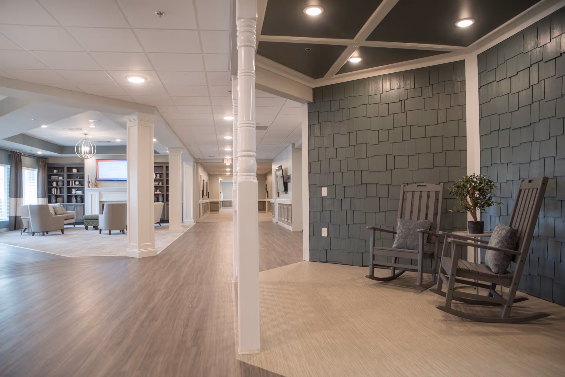 Interior view of a senior living facility showing a spacious hallway with wood flooring and a seating area with two wooden rocking chairs and a small table with a potted plant. In the background, there is a living room area with armchairs, a fireplace, built-in shelves, and a wall-mounted TV.