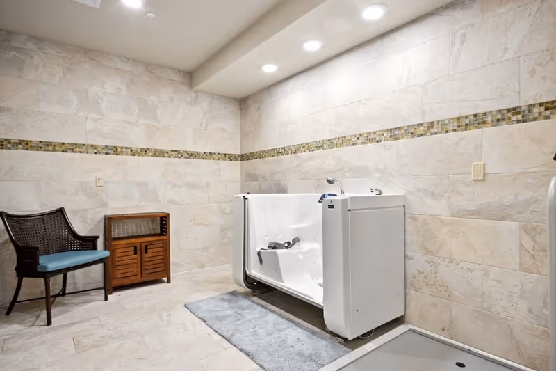 A spacious bathroom with beige tiled walls and floor, featuring a white walk-in bathtub with safety handles and a blue cushioned wicker chair next to a small wooden cabinet. The room is well-lit with recessed ceiling lights and has a decorative horizontal tile border.