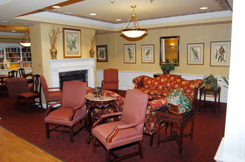 Carpeted common living area with upholstered chairs and a patterned sofa around small tables, a fireplace, framed bird prints, and bookshelves in the background.
