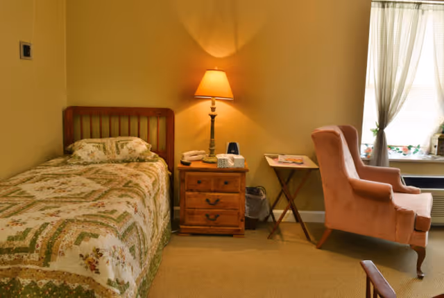 A cozy bedroom with a single bed covered in a patterned quilt, a wooden nightstand with a lamp, telephone, and tissues, a pink upholstered armchair near a window with sheer curtains, and a small folding table with magazines.
