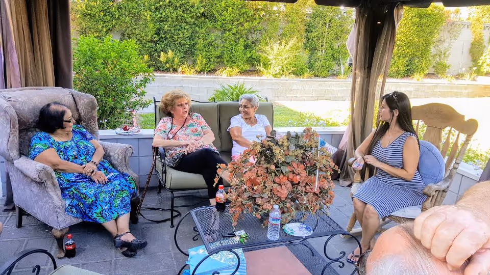 Four women sitting and chatting in an outdoor covered patio area with cushioned chairs and a metal table with a floral centerpiece, water bottles, and a soda bottle. Green bushes and a stone wall are visible in the background.
