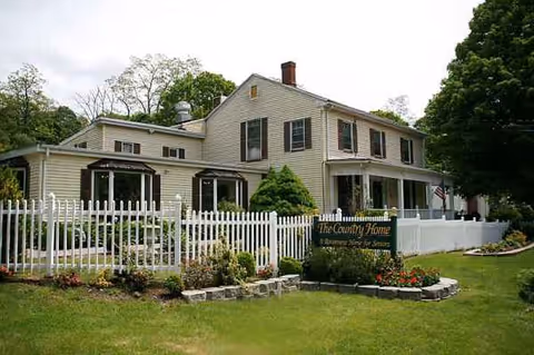 Front view of a two-story senior living house with white picket fence, garden, and sign