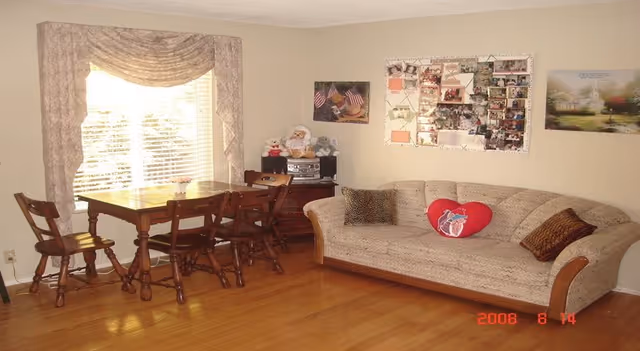 A cozy living room with a wooden floor, a beige patterned sofa with three pillows including a red heart-shaped one, a wooden dining table with four chairs near a window with blinds and floral curtains, and a small wooden cabinet with a stereo and stuffed animals. The walls have several framed pictures and a bulletin board with photos and notes.