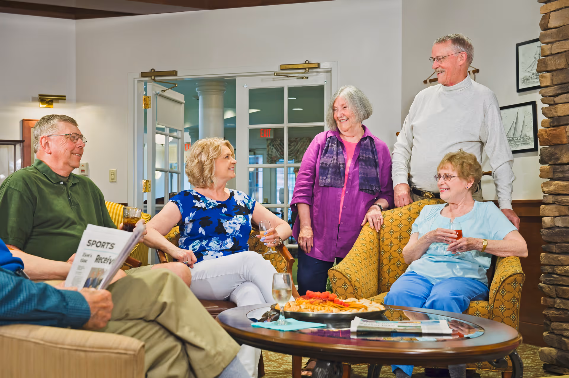 A group of five elderly people socializing in a cozy living room area. Two women and one man are seated on comfortable chairs and a sofa, holding drinks, while another woman and man stand nearby, all smiling and engaging in conversation. A round coffee table with snacks and newspapers is in the center, and the room features warm lighting and a stone fireplace.