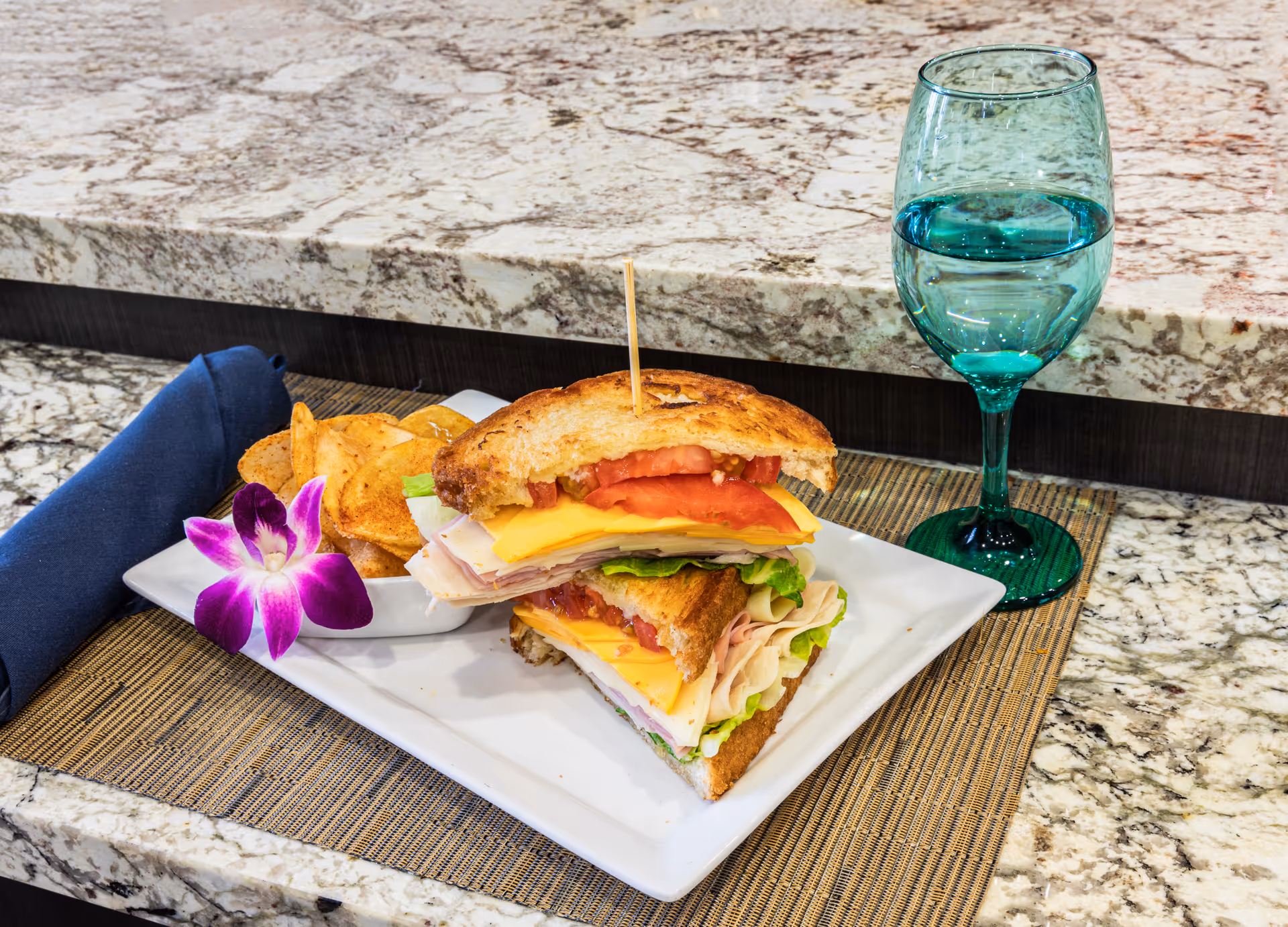 A club sandwich with layers of turkey, cheese, lettuce, and tomato on toasted bread, served on a white rectangular plate with a side of potato chips and a purple orchid flower. A blue cloth napkin is rolled up beside the plate, and a green-tinted glass of water is placed on a granite countertop.