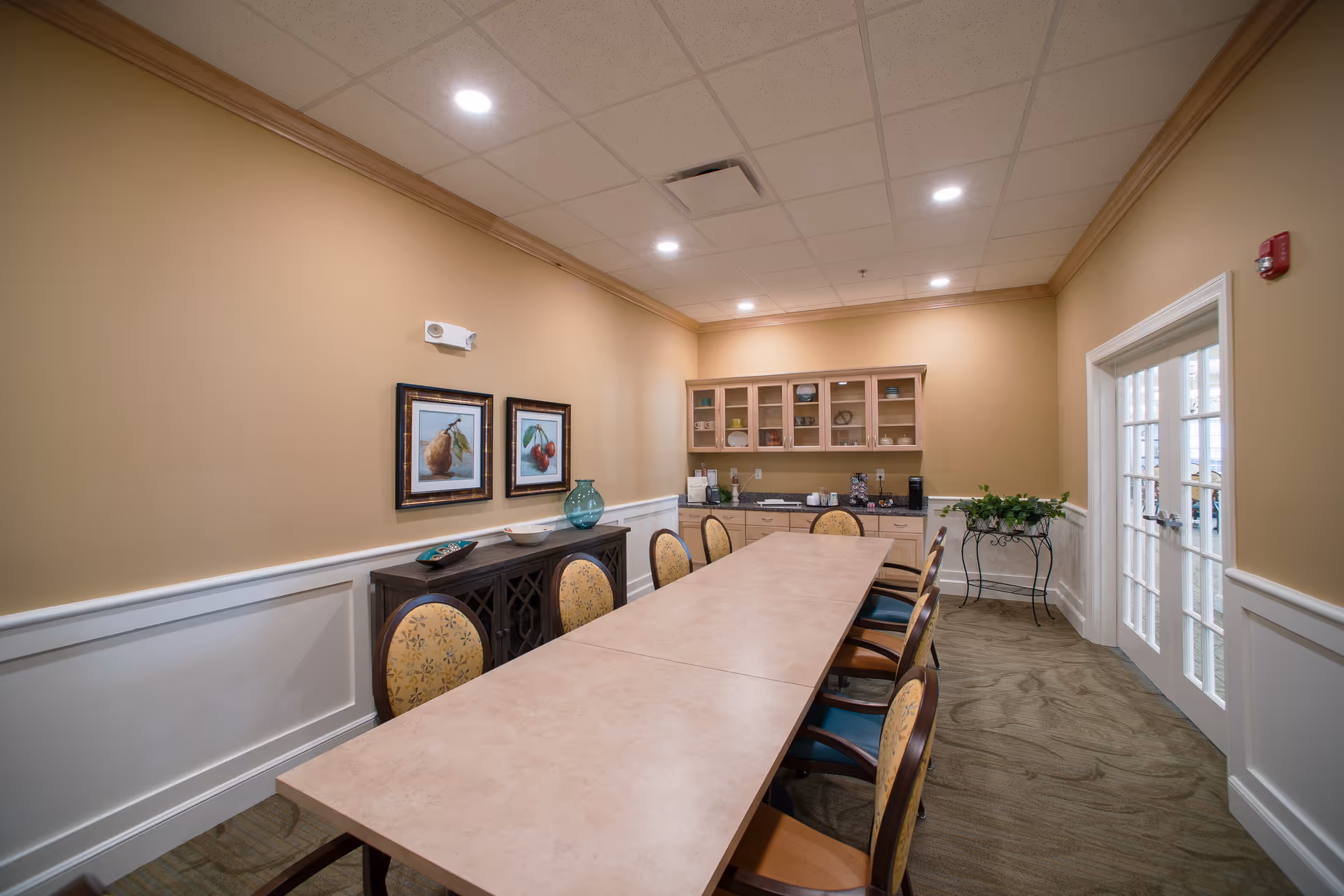 Long dining/meeting room with a central rectangular table surrounded by chairs, cabinets and framed artwork, and French doors to the side.