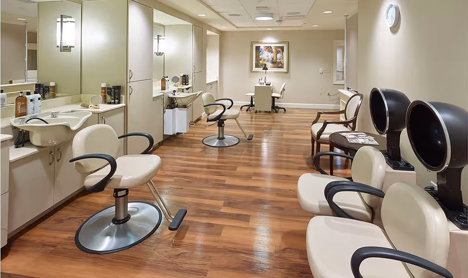 Interior view of a salon area with beige styling chairs, hair washing sinks, hair dryers, and a small desk with chairs in the background. The room has wooden flooring, light-colored walls, and a framed picture on the far wall.