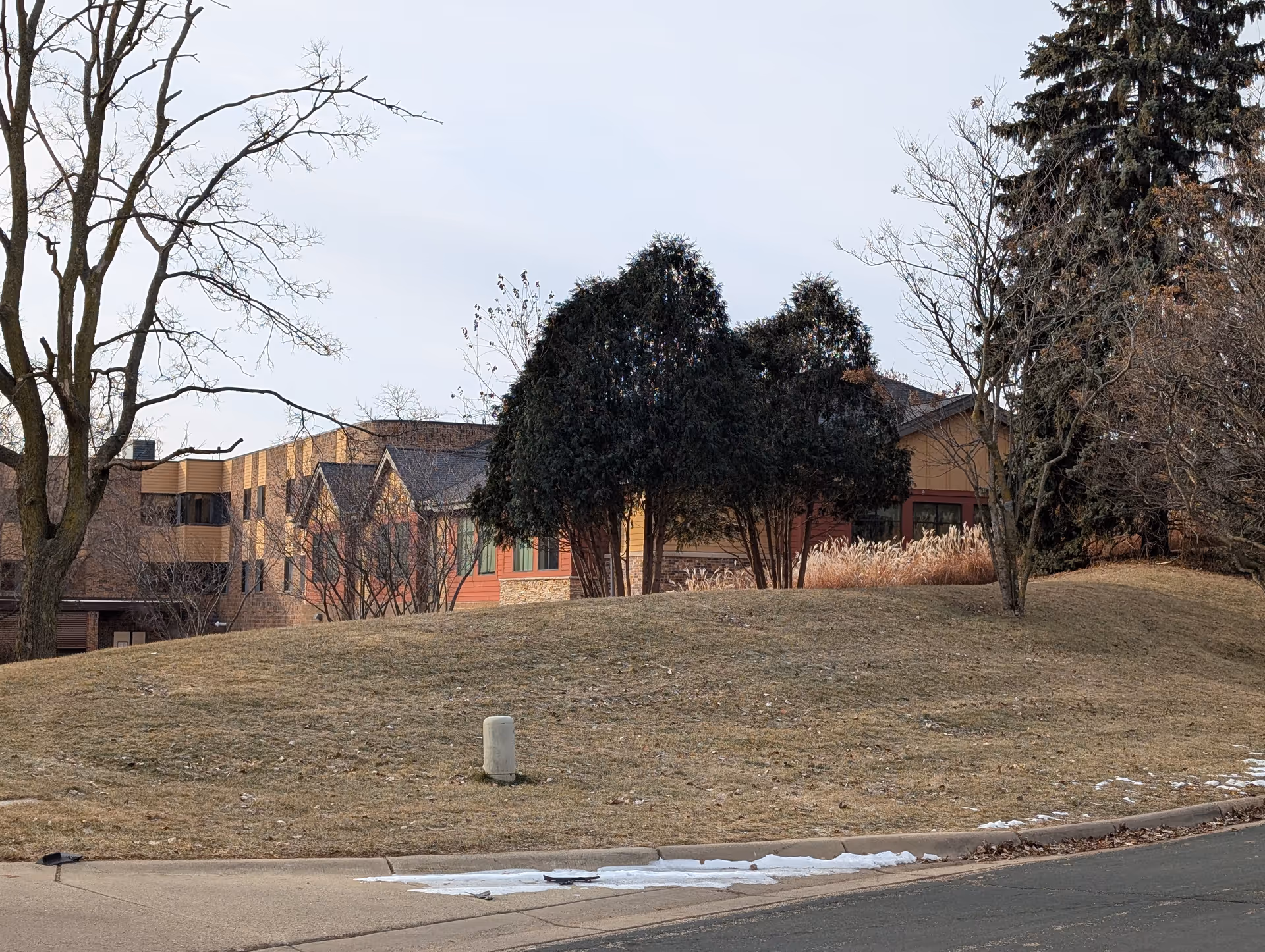 View of a senior living facility building partially obscured by trees and bushes on a grassy hill with a sidewalk and street in the foreground. The building has a mix of brick and siding exterior with multiple windows and a sloped roof. Some patches of snow are visible on the grass near the sidewalk.