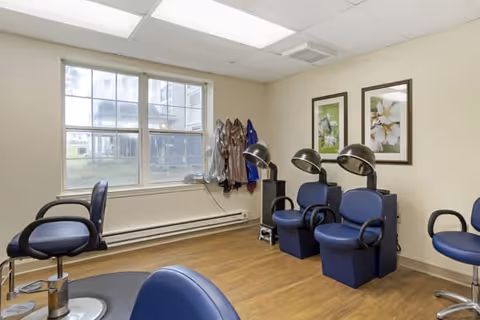 Interior view of a hair salon area in a senior living facility with blue salon chairs, hair drying stations, a large window, and framed floral artwork on the wall.