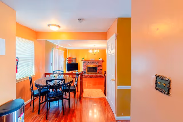 Interior view of a living and dining area with orange walls and polished wooden floors. The dining area has a round table with six black metal chairs. Beyond the dining area is a living room with a brick fireplace, a TV on a stand, and a chandelier hanging from the ceiling.