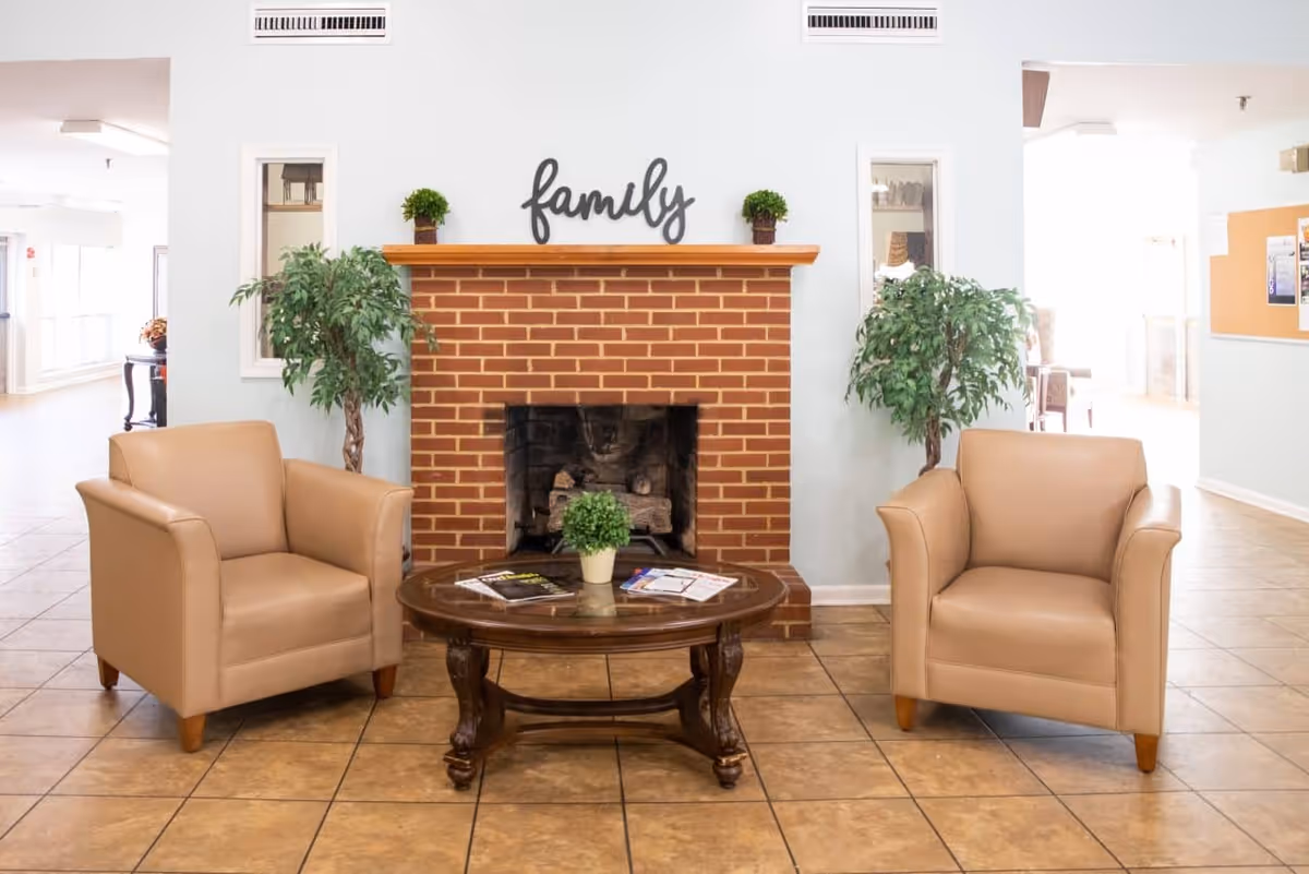 Two beige armchairs flank a brick fireplace with a coffee table and potted plants in a sitting area.