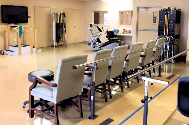 A rehabilitation room with a row of chairs lined up along parallel bars used for walking exercises. The room also contains exercise equipment including a stationary bike, a TV mounted on the wall, and various therapy tools and supplies organized on shelves and racks.