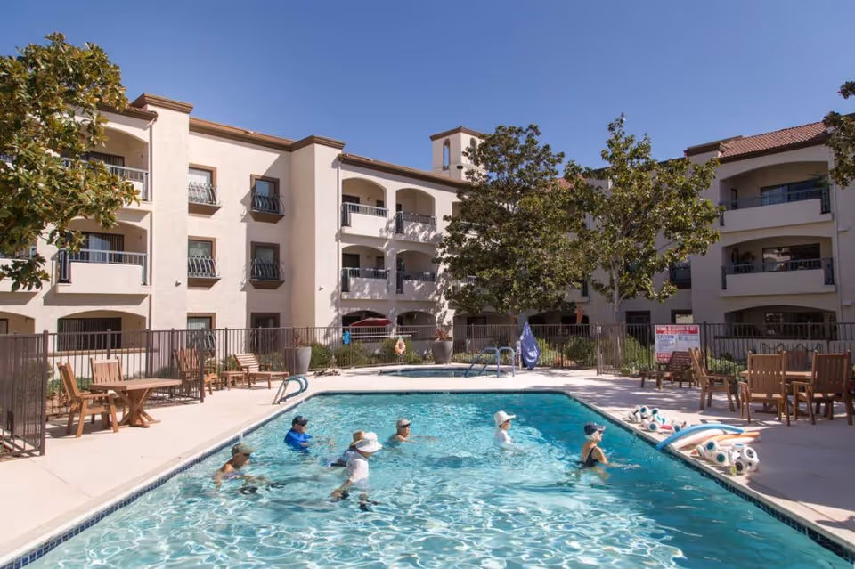 Outdoor swimming pool at Casa de las Campanas with several elderly people participating in a water exercise class. The pool is surrounded by a concrete deck with wooden tables and chairs, and the multi-story residential building with balconies is visible in the background under a clear blue sky.
