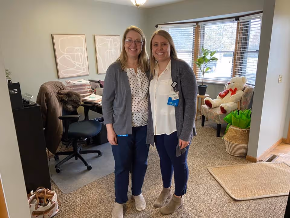Two women standing and smiling inside an office area with a desk, chair, and paperwork. Behind them is a window with blinds, a potted plant, a chair with a large teddy bear holding a heart, and a basket with green umbrellas.