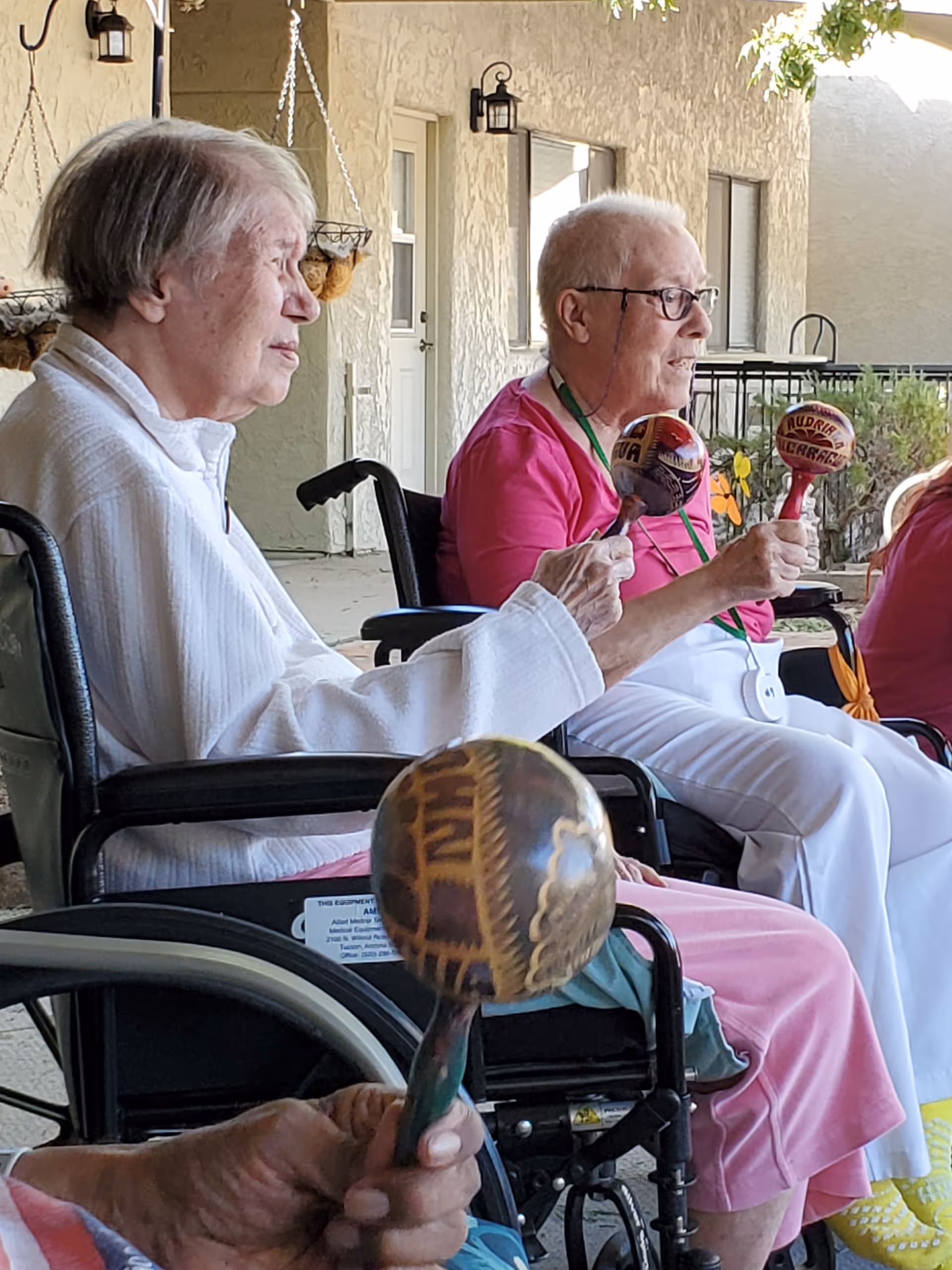 Three elderly individuals sitting in wheelchairs on a covered outdoor patio, each holding and shaking a decorative maraca. The background shows textured walls, windows, doors, and hanging planters.
