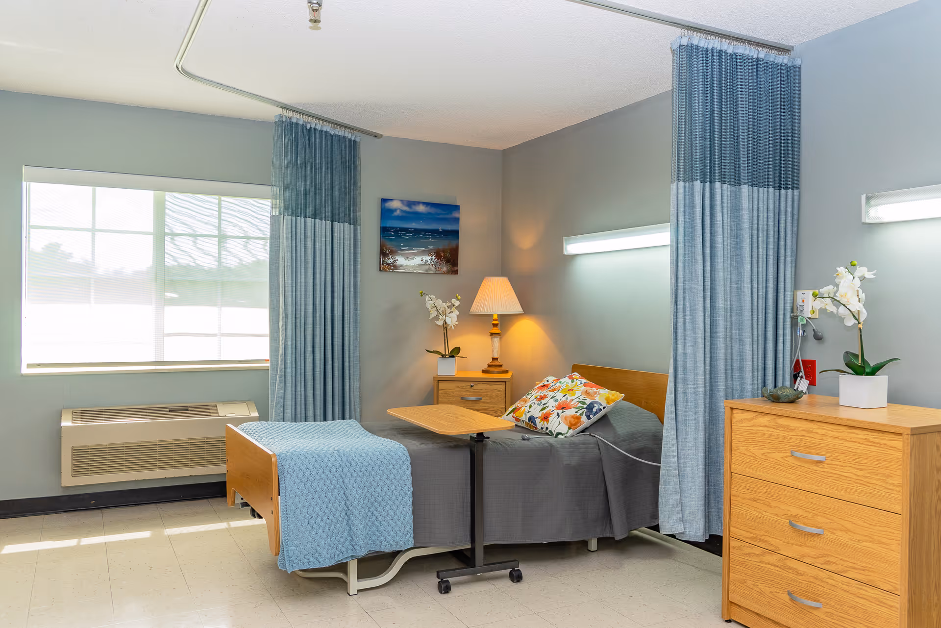 A nursing facility bedroom with a single bed covered in a gray blanket and a blue knitted throw. The bed has a floral pillow and an adjustable overbed table. There are blue privacy curtains on either side of the bed. A wooden nightstand with a lamp and a white orchid plant is next to the bed. A wooden dresser with another white orchid plant is on the right side. A window with blinds and an air conditioning unit is on the left wall. A framed picture of a beach scene hangs on the wall above the nightstand.