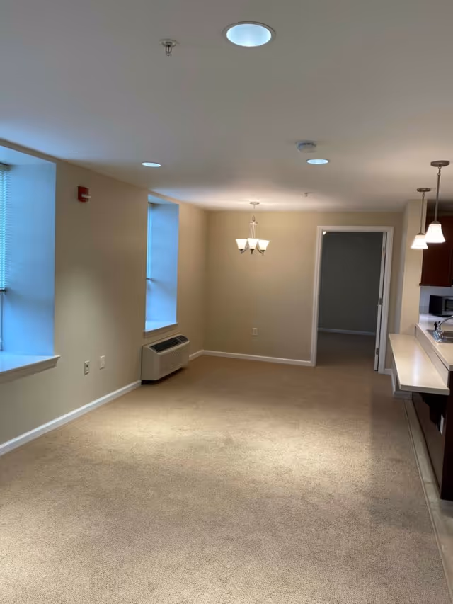 Empty interior room with beige carpet and walls, two windows with blinds, a wall-mounted air conditioning unit, ceiling lights including a chandelier, and a kitchen counter with hanging pendant lights on the right side.