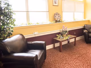 A cozy sitting area with two dark leather armchairs facing each other, a small wooden table with a floral arrangement between them, and large windows with blinds letting in natural light. The walls are painted yellow and maroon, and there is a patterned carpet on the floor.