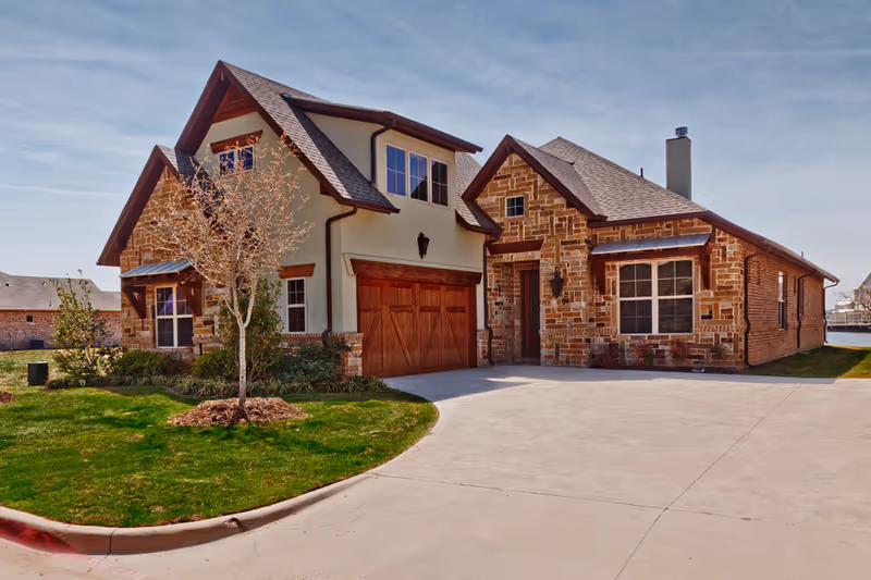 Exterior view of a residential-style building with a stone and stucco facade, a two-car wooden garage door, and a small tree planted in a grassy area near the driveway under a clear sky.