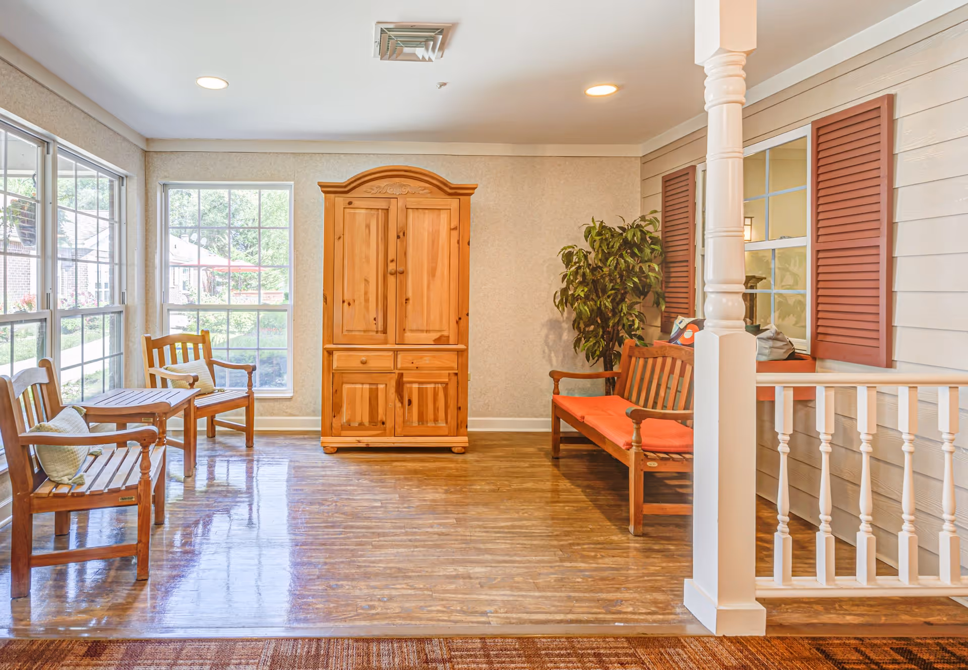 A bright indoor seating area with wooden furniture including two chairs with cushions and a small table on the left, a wooden cabinet in the center against the wall, and a wooden bench with an orange cushion on the right. The room has large windows letting in natural light, a potted plant next to the bench, and a white railing with a decorative post on the right side.