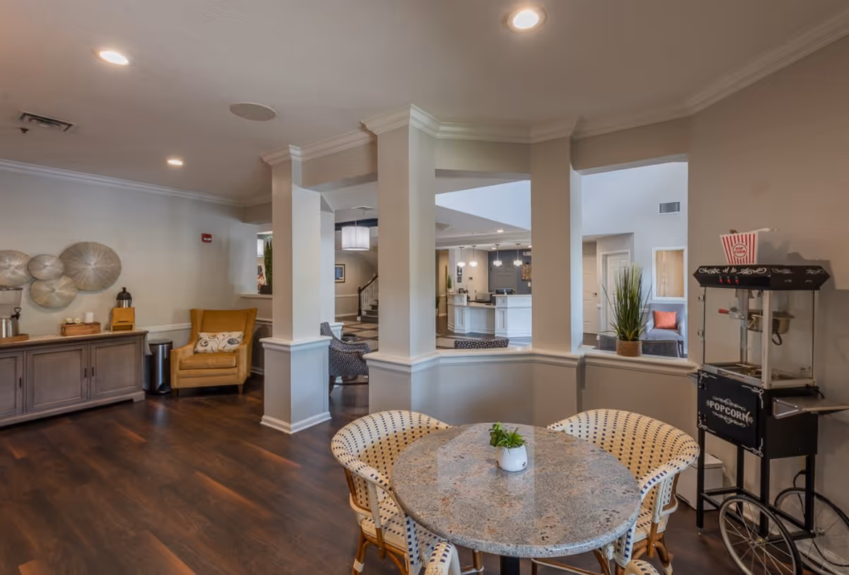 Interior view of a senior living facility lounge area with a round table and four wicker chairs, a popcorn machine on the right, a yellow armchair and a wooden cabinet on the left, and a reception desk visible in the background.