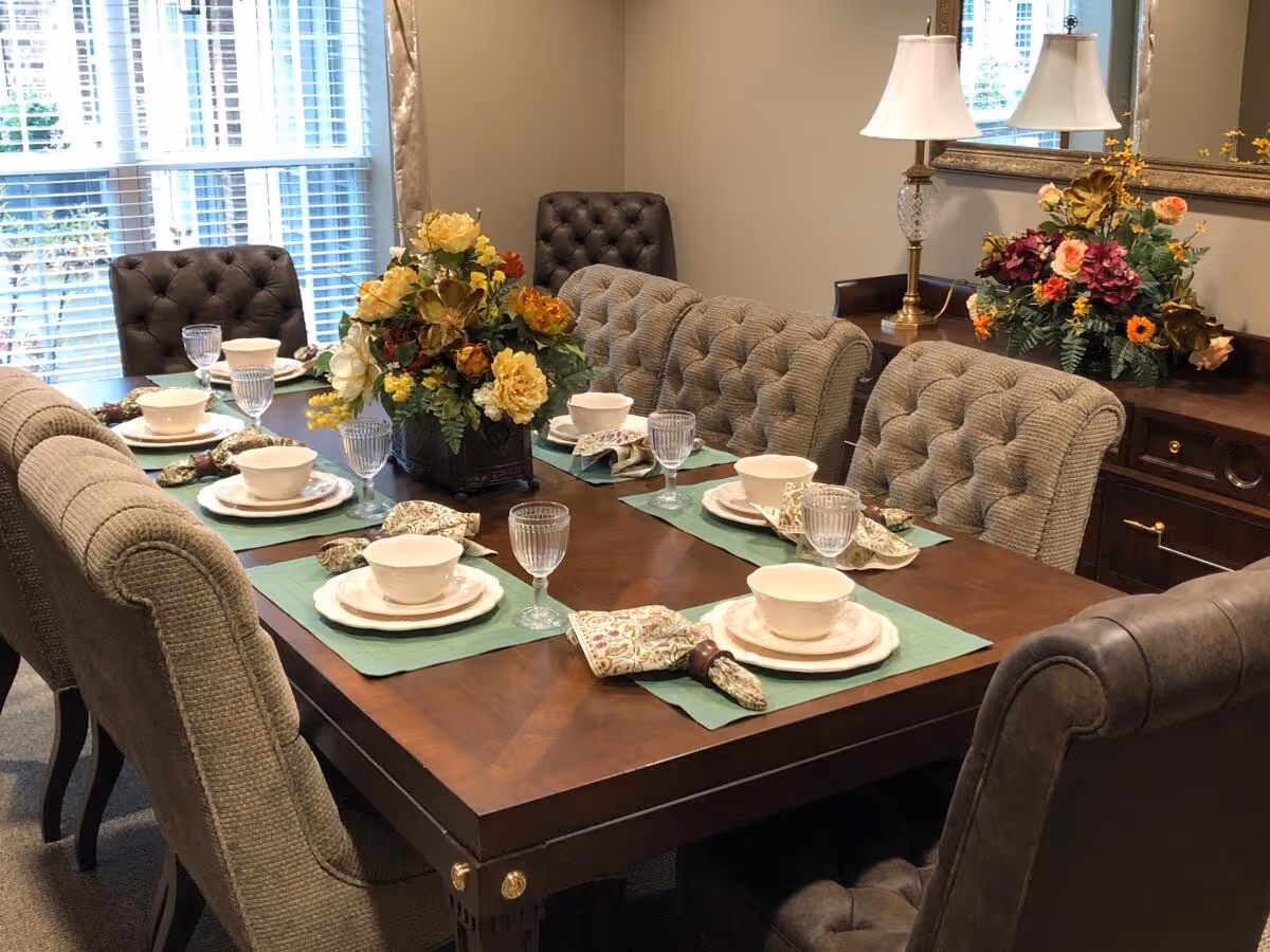 A formal dining room with a dark wooden table set for six people. The table is adorned with green placemats, white bowls, plates, glassware, and patterned napkins with napkin rings. There is a large floral centerpiece in the middle of the table. The room has tufted upholstered chairs around the table, a sideboard with a lamp and another floral arrangement, and a large window with white blinds letting in natural light.