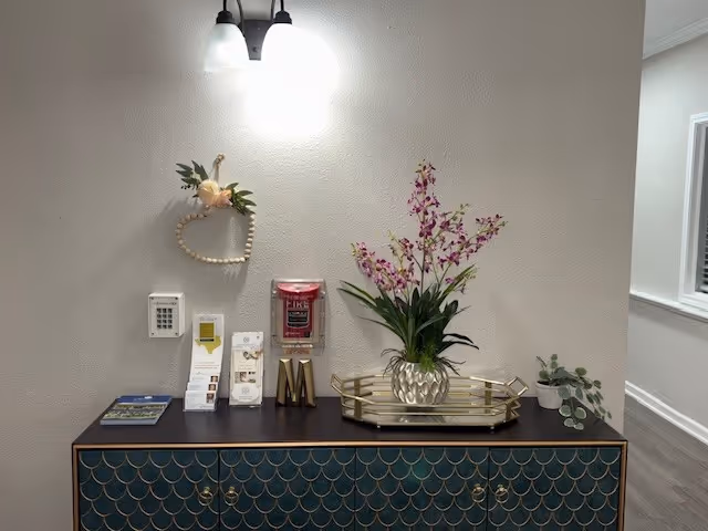 A decorative console table with a dark top and blue-green scale-patterned front is placed against a light gray wall. On the table, there is a gold tray holding a vase with purple flowers, a small potted plant, brochures, and a fire alarm pull station mounted on the wall above. A heart-shaped wall decoration with flowers hangs next to a wall light fixture with two bulbs.