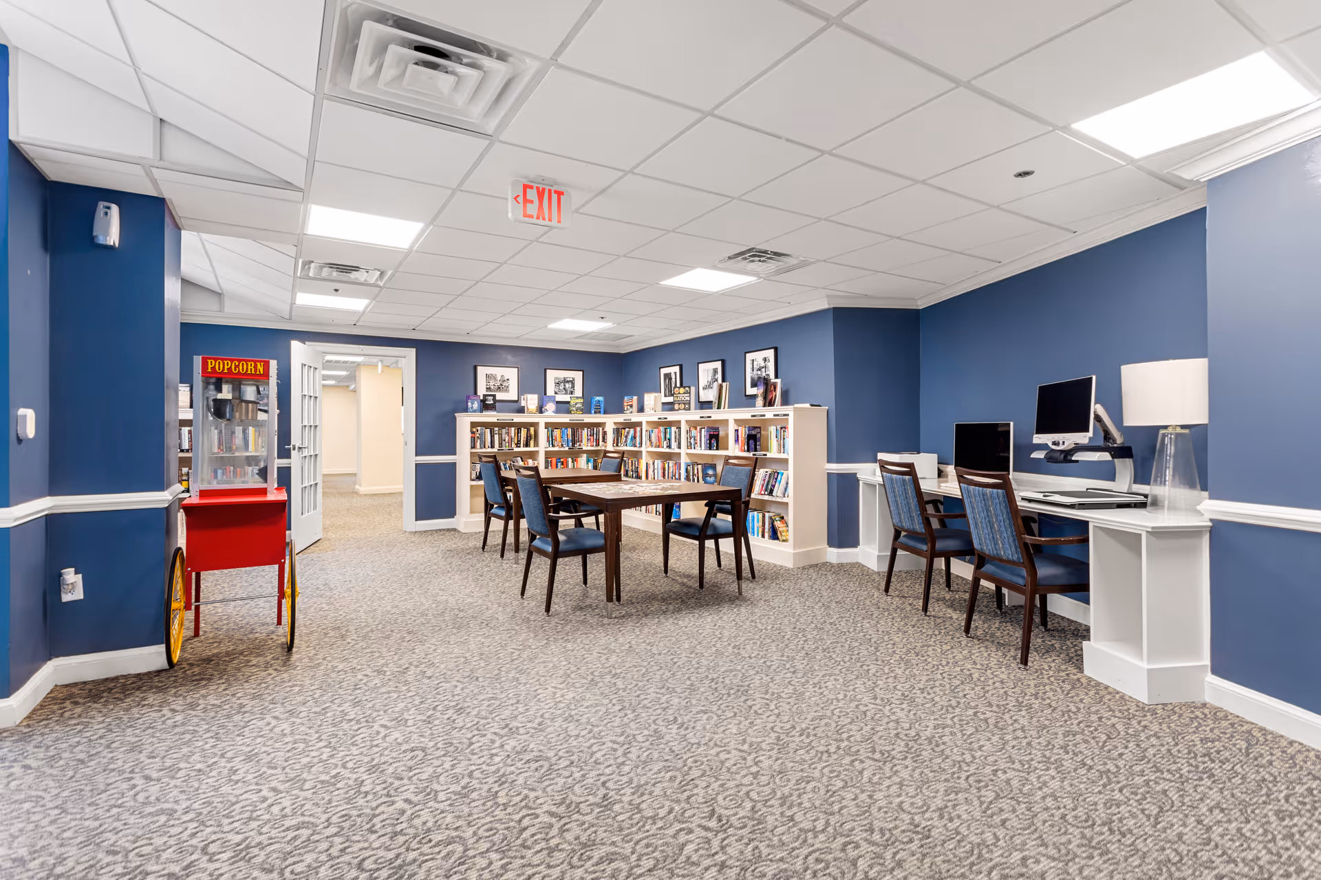 A community room with blue walls and patterned carpet featuring a popcorn machine on the left, a table with four chairs in the center, bookshelves filled with books along the back wall, and a computer workstation with two chairs and a lamp on the right side. The ceiling has recessed lighting and an exit sign is visible above a doorway.