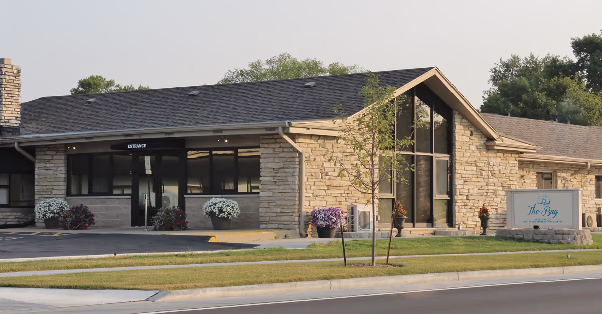 Exterior front of a one-story stone-clad senior care building with a glass entrance, potted flowers, and a sign reading 'The Bay'.