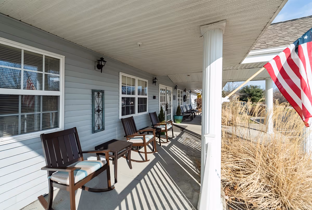 Covered porch area at Cedar Creek of Bloomington Assisted Living with several wooden rocking chairs and small tables along the wall. The porch has white columns and overlooks a garden area with tall grasses. An American flag is mounted on one of the columns.