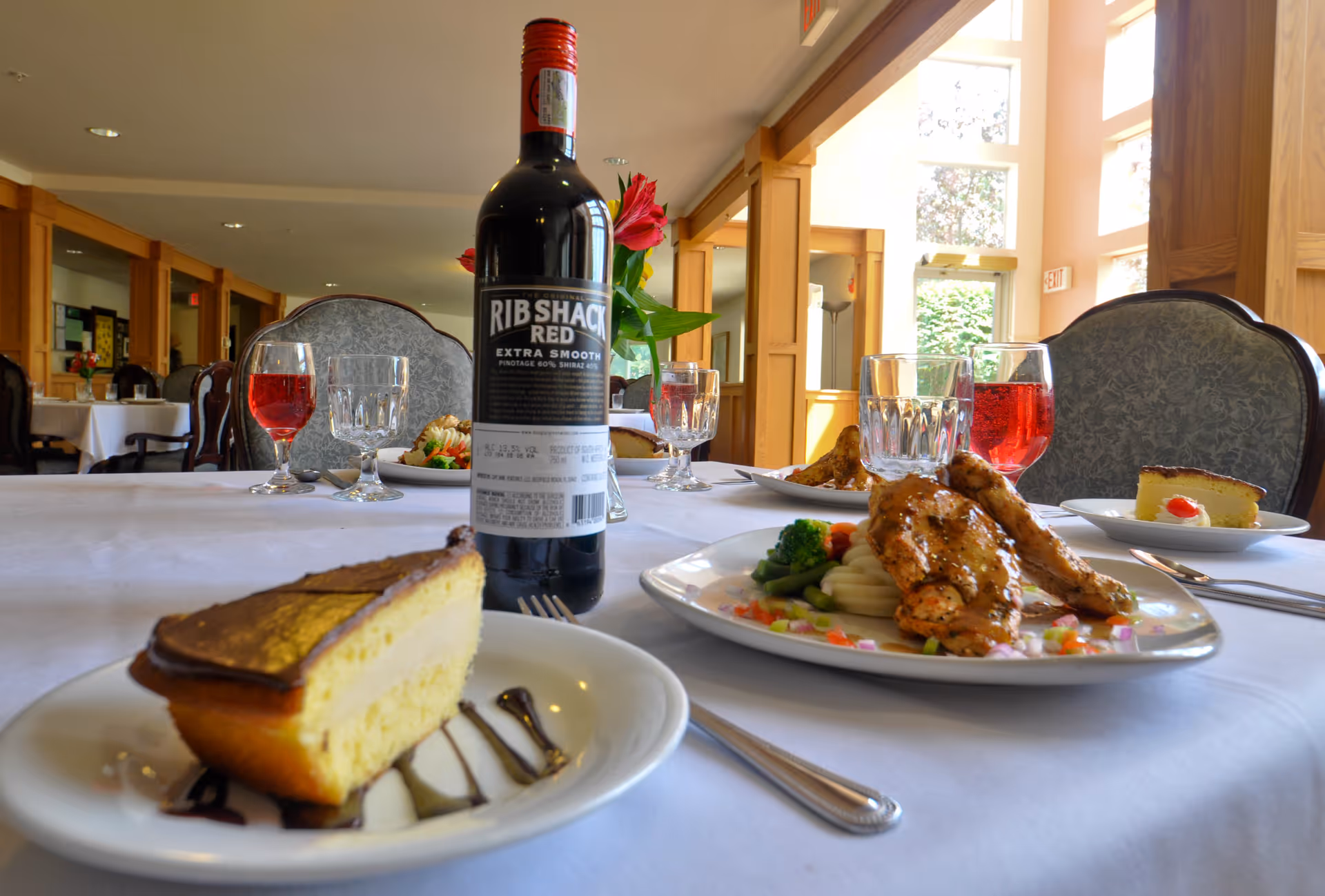 A dining table set with plates of food including a slice of cake and a grilled chicken dish with vegetables, glasses of water and red beverage, and a bottle of Rib Shack Red wine in a well-lit dining room with wooden paneling and upholstered chairs.