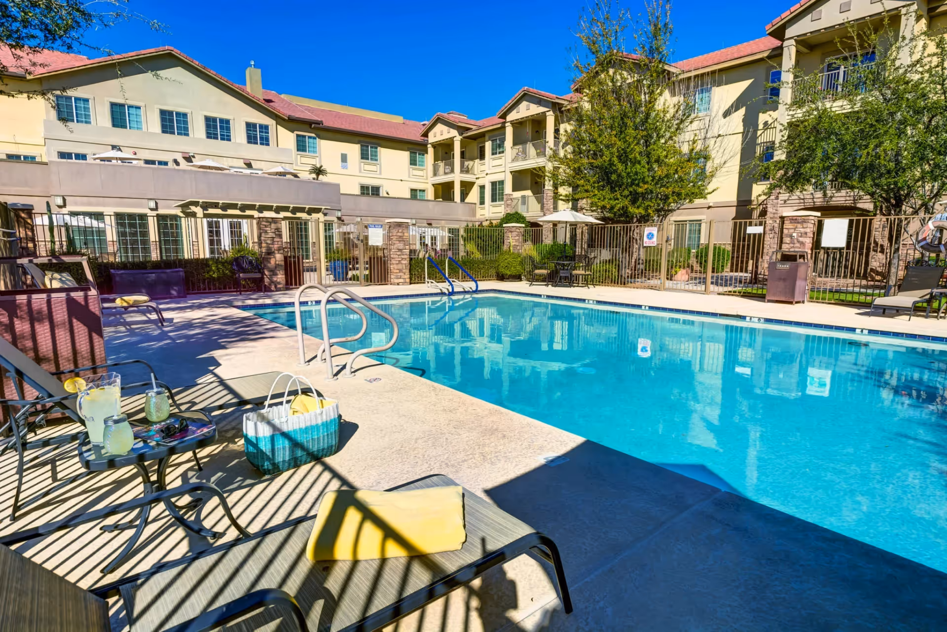 Outdoor swimming pool area at a senior living facility with lounge chairs, a small table with drinks, and a basket. The pool is surrounded by a fence and multi-story residential buildings with balconies and trees under a clear blue sky.