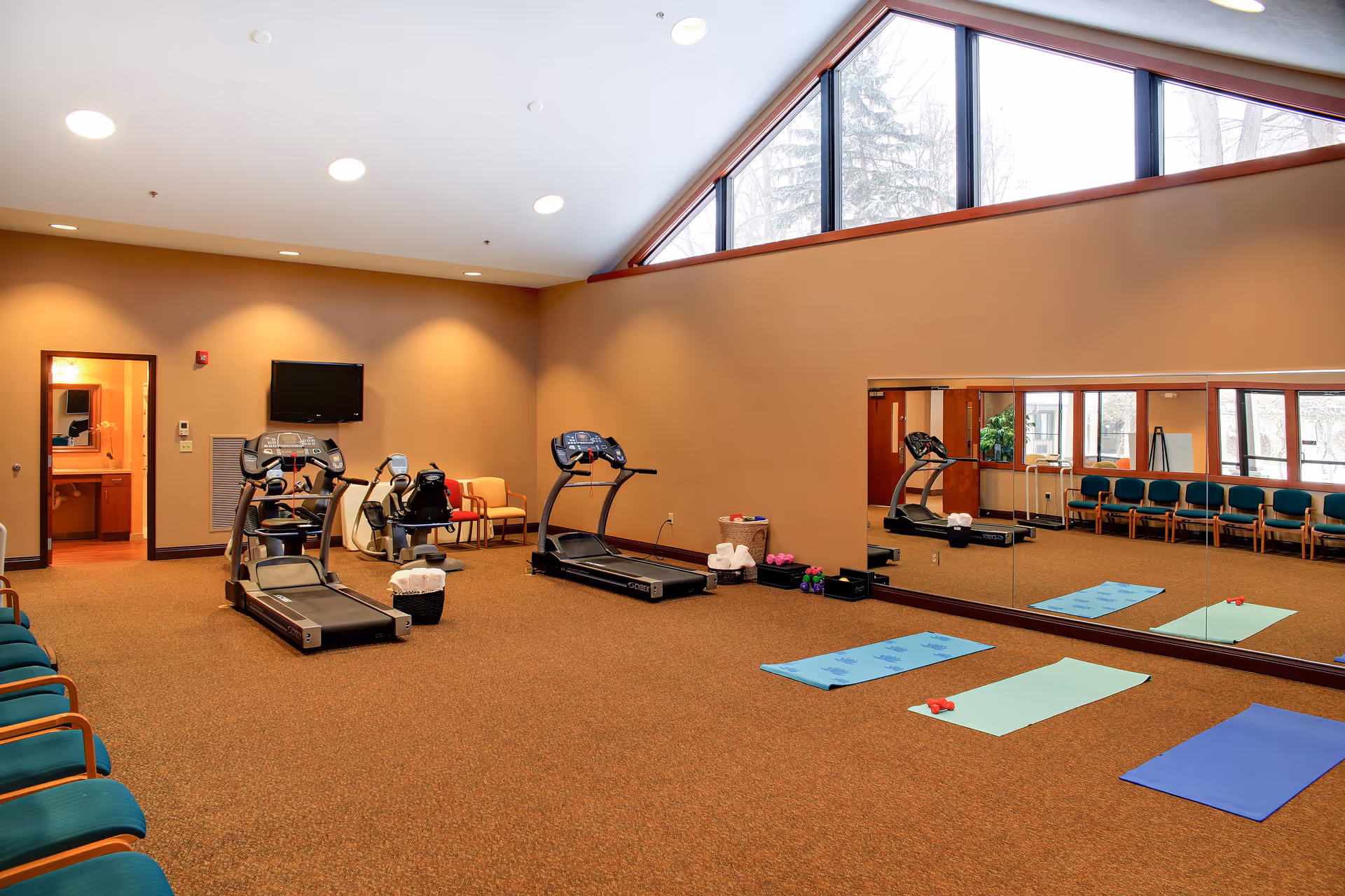 Bright exercise room with treadmills, yoga mats, large mirror, and seating under a high windowed ceiling.