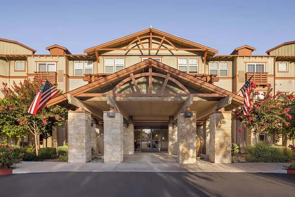 Front entrance of a multi-story senior living building with a covered porte-cochere, stone pillars, American flags, and landscaped beds.