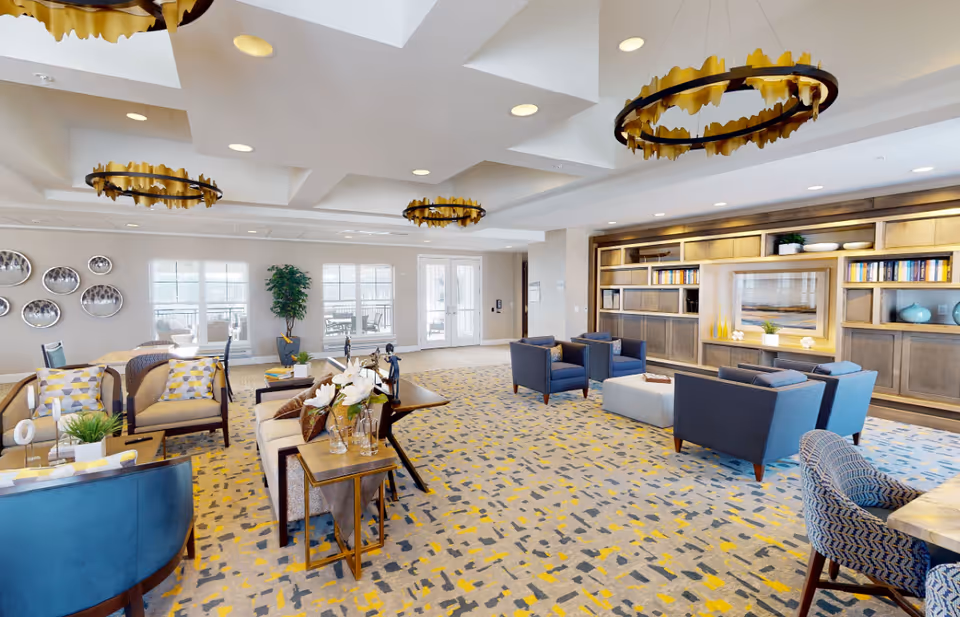 A spacious and well-lit senior living common area with modern furniture including armchairs, sofas, and tables. The room features a patterned carpet with yellow, gray, and blue accents, decorative wall art, large windows letting in natural light, and built-in wooden shelves with books and decorative items. Three unique circular gold chandeliers hang from the ceiling.