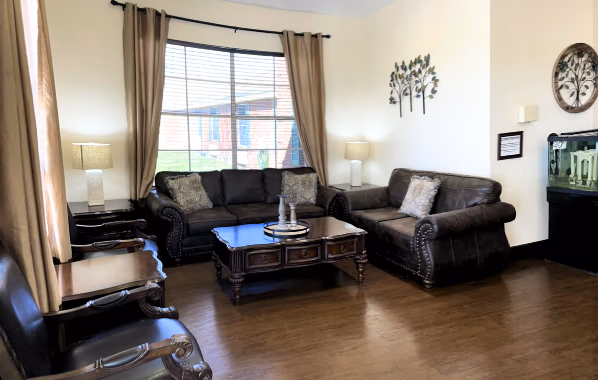 A cozy living room with two dark brown leather sofas, a wooden coffee table with decorative items, two matching table lamps on side tables, beige curtains framing a large window, wooden chairs with armrests, and wall decorations including metal tree art and a round frame. There is also a fish tank in the corner.