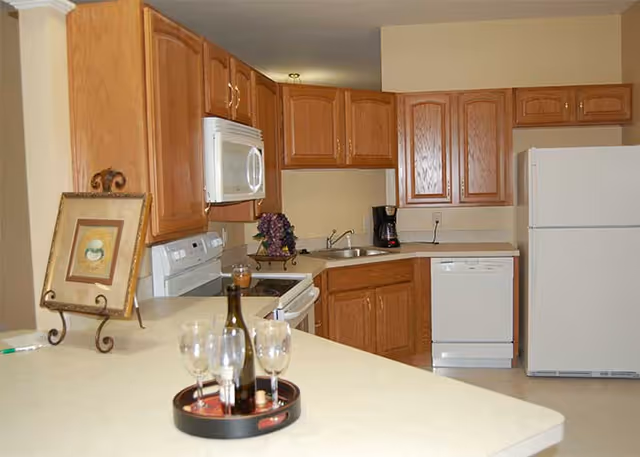 A clean kitchen with oak cabinets and white appliances, a countertop island holding a wine bottle and glasses, and a framed picture on a stand.
