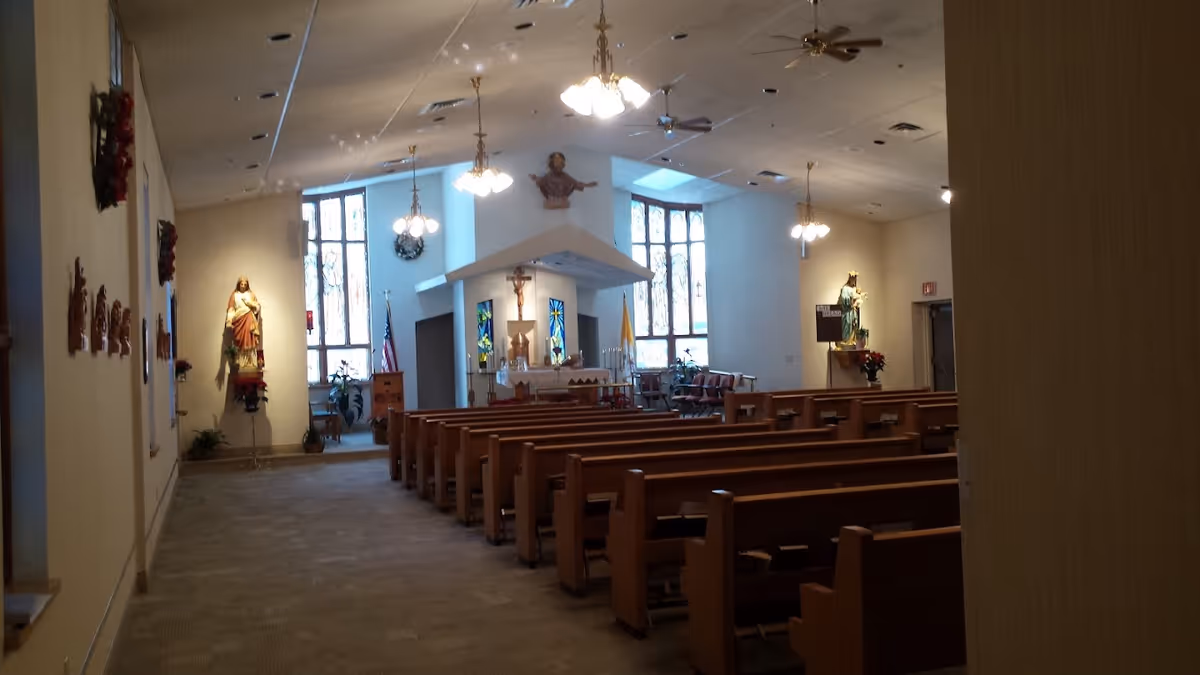 Interior view of a chapel with wooden pews facing an altar, stained glass windows, and religious statues.