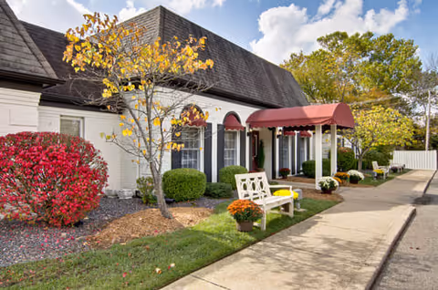 Exterior view of a single-story building with a dark shingled roof and white siding. The entrance features a red awning and white columns. There are benches, potted plants, and landscaped bushes and trees with autumn foliage along the sidewalk leading to the entrance.
