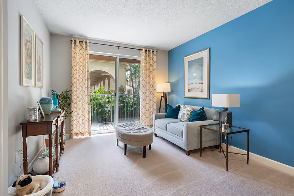 A bright living room with a light gray sofa adorned with blue and patterned cushions against a blue accent wall. There is a glass side table with a lamp, a floor lamp, and a framed picture on the wall. A tufted oval ottoman is placed in front of the sofa. On the opposite side, a wooden console table holds decorative items and plants. The room has beige carpet flooring and sliding glass doors with patterned curtains leading to an outdoor balcony with greenery visible outside.