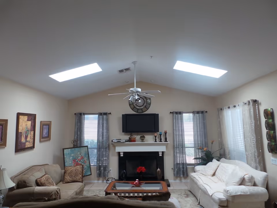 Bright living room with sofas, a coffee table, a fireplace and wall-mounted TV under skylights and a ceiling fan.