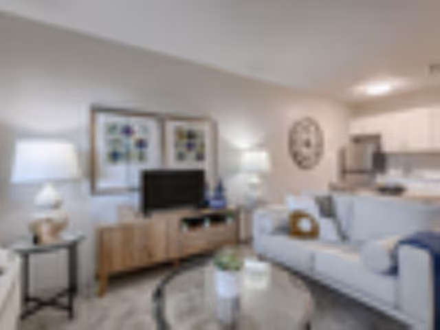 Bright furnished living room with a sofa, coffee table, TV on a media console, and an open kitchen in the background.