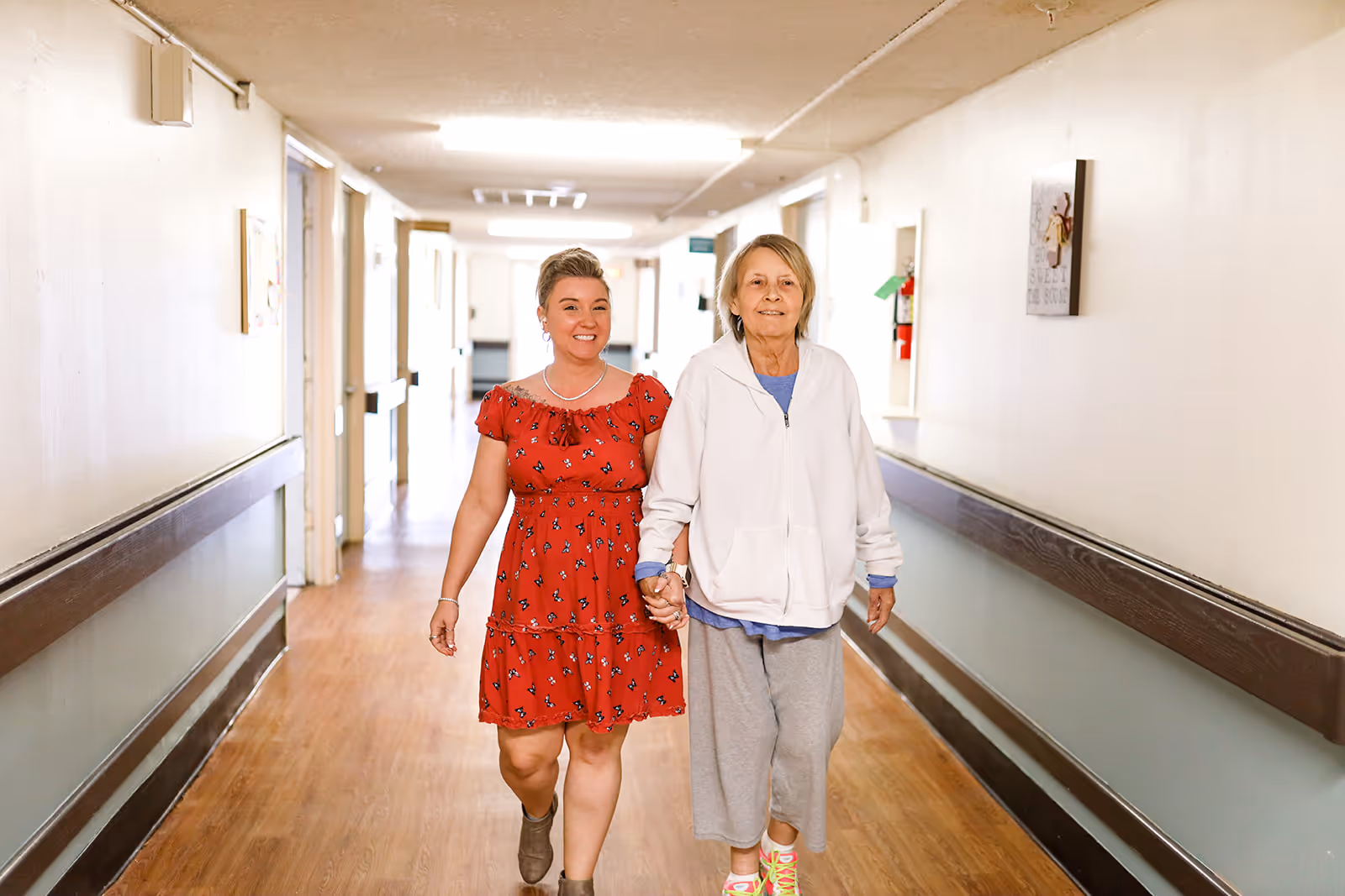 Two women walking hand in hand down a well-lit hallway in a healthcare facility. One woman is elderly, wearing a white jacket and gray pants, while the other is younger, wearing a red dress with a floral pattern. The hallway has wooden floors, handrails on both sides, and doors leading to rooms.