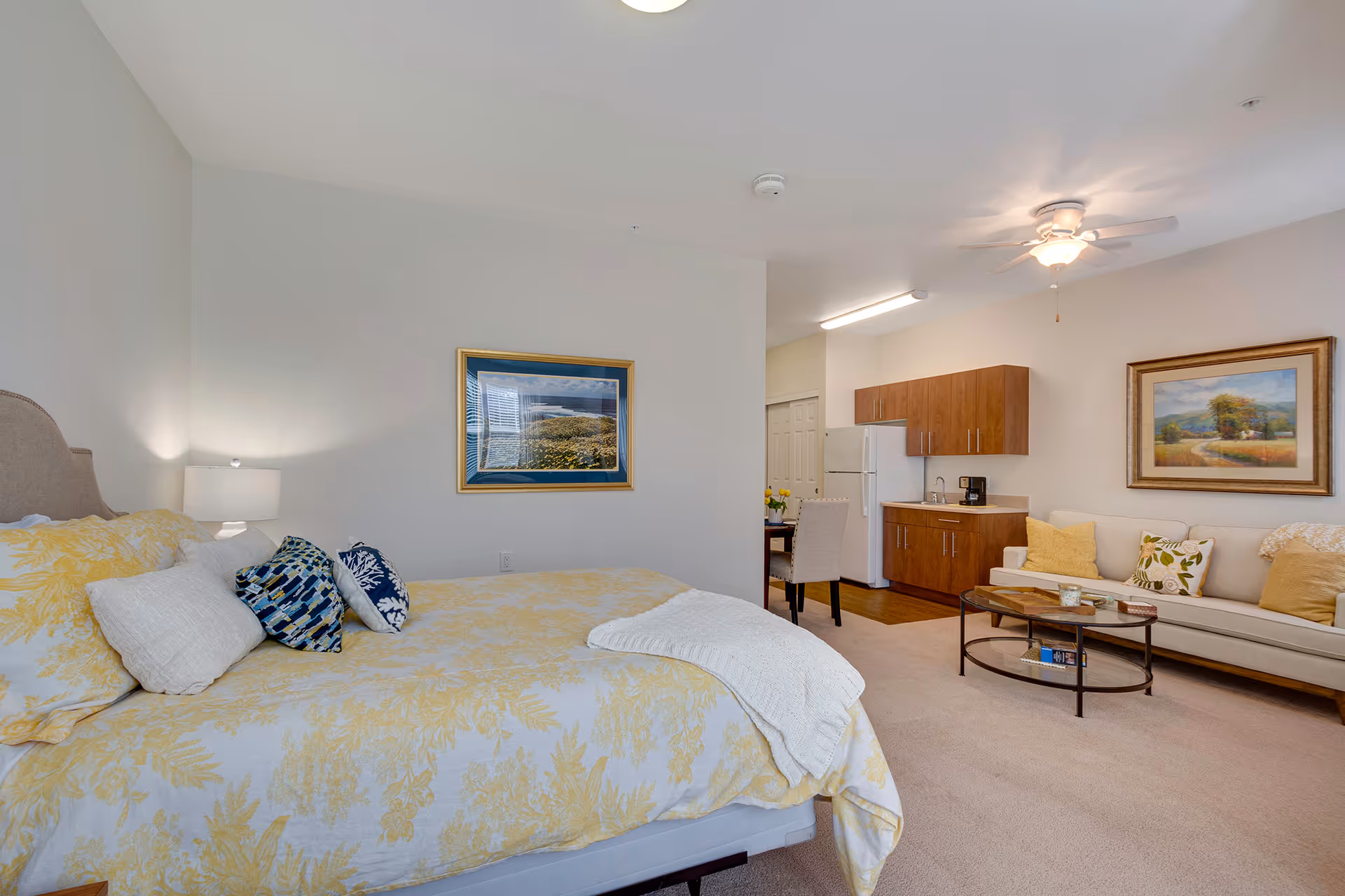 Bright studio apartment interior showing a bed with yellow bedding, a kitchenette, and a seating area with a sofa and coffee table.